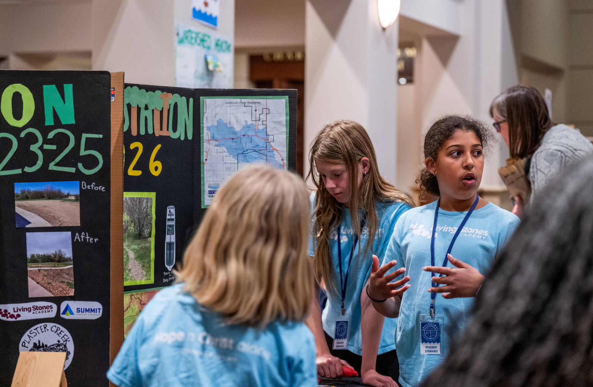 three 6th grade students in light blue T-shirts that say "living stones academy" on them, students talking and discussing their posters to an audience off-camera